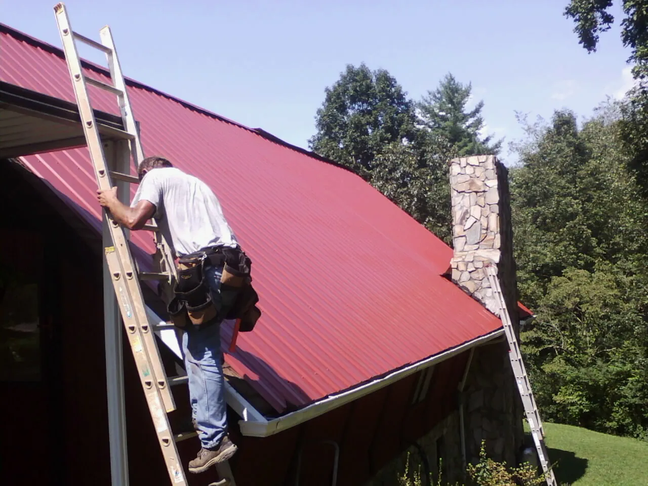 Roofer on a ladder beside a red metal roof and stone chimney — Soffit & Fascia Repair by Roofing Brothers in Eastford